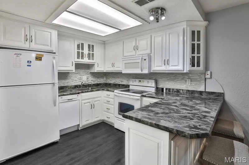 Kitchen with white appliances, decorative backsplash, a peninsula, white cabinetry, and glass insert cabinets — 1380 Heritage Landing #206, St Charles, MO 63303