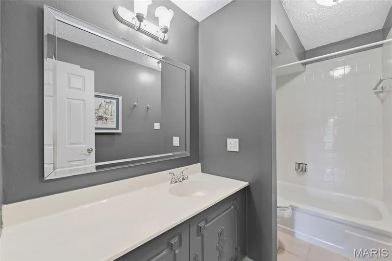 Full-length mirror with beveled edges, vanity with white countertop, chrome faucet, white tile shower with tub, and wall-mounted light fixture — 14443 Bantry Lane #16, Chesterfield, MO 63017
