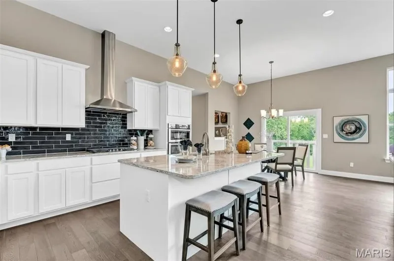 Kitchen with decorative backsplash, a breakfast bar, an island with sink, wall chimney range hood, and light stone counters — 1 TBB Monaco @Willow Grove, Dardenne Prairie, MO 63368