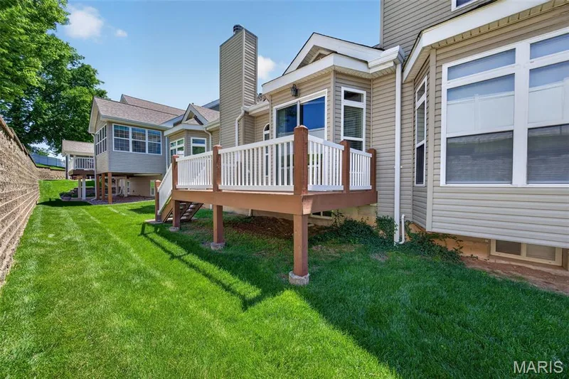 Rear exterior featuring a spacious deck with white railings, tan vinyl siding, multiple windows, and a retaining wall — 216 Woodland Place Court, St Charles, MO 63303