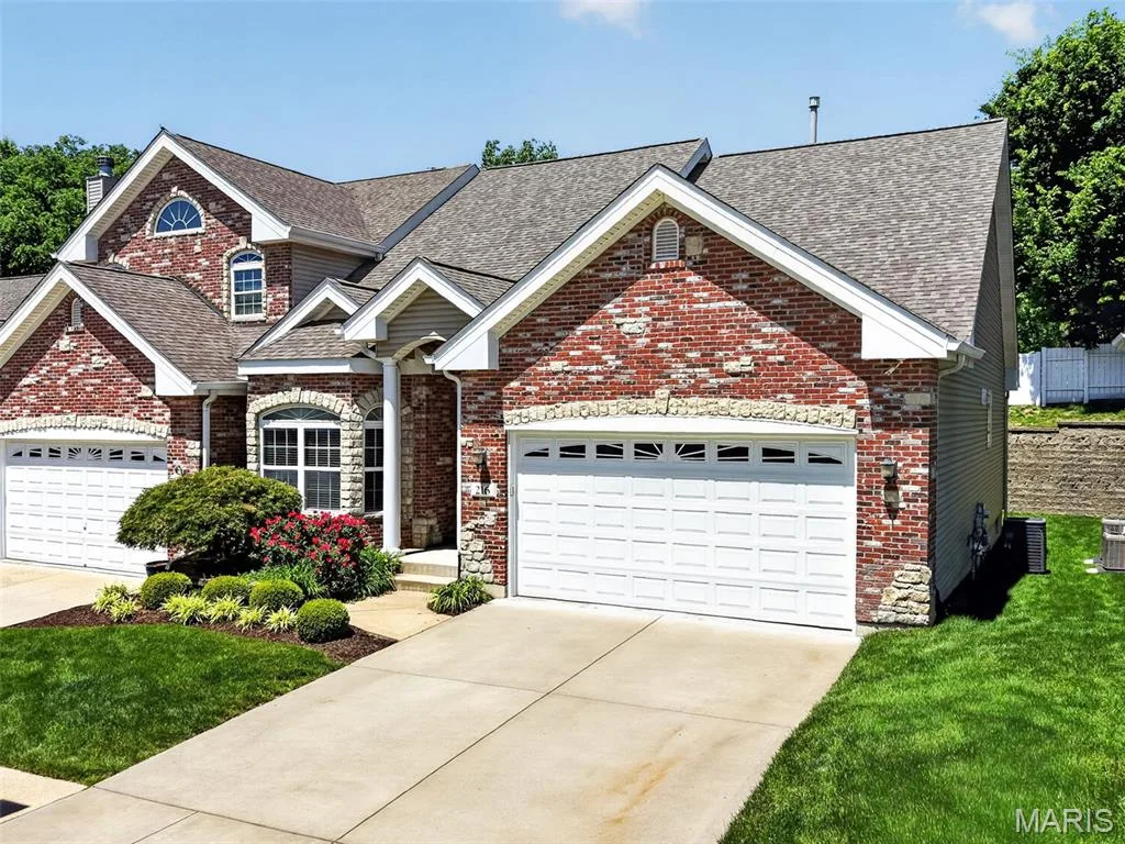 Red brick exterior with stone accents, two-car garage, manicured front landscaping, and a concrete driveway — 216 Woodland Place Court, St Charles, MO 63303