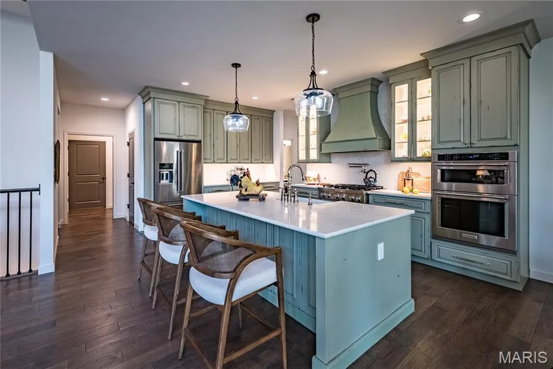 This spacious kitchen features hardwood flooring, an expansive island with seating, stainless steel appliances, and custom cabinetry with illuminated glass-front uppers — 150 Delcreek Estates Drive, Foristell, MO 63348