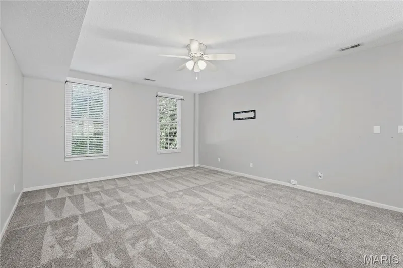 Expansive room featuring two windows, patterned carpet, a ceiling fan, and light gray walls — 3245 Domain Street, St Charles, MO 63301