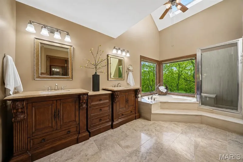 Dual wood-finish vanities with light-toned countertops, a built-in corner soaking tub, a separate glass-enclosed shower, and a vaulted ceiling with skylight and ceiling fan — 1553 Ridge Road, Wildwood, MO 63021