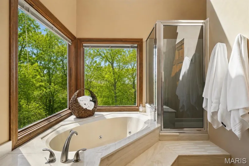 Corner jetted bathtub with a brushed nickel faucet, set within a light-toned stone surround, illuminated by two large windows with wood-finish trim, and an enclosed shower with a glass door — 1553 Ridge Road, Wildwood, MO 63021