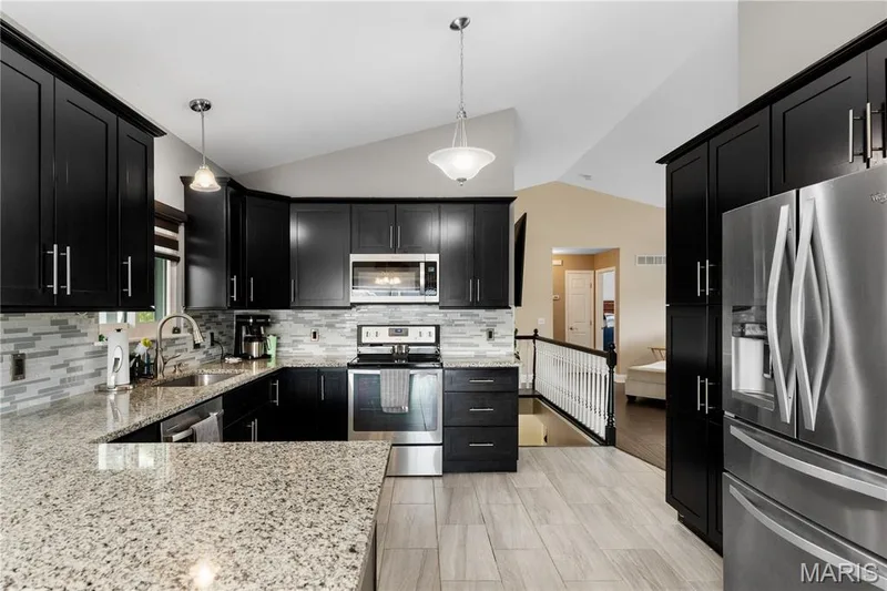 Kitchen with dark cabinets, appliances with stainless steel finishes, light stone counters, lofted ceiling, and hanging light fixtures — 10107 Greenbriar Court, Foristell, MO 63348