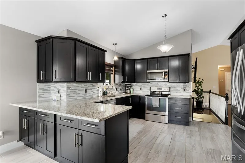 Kitchen with stainless steel appliances, hanging light fixtures, light stone countertops, dark cabinets, and decorative backsplash — 10107 Greenbriar Court, Foristell, MO 63348