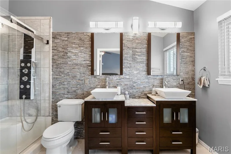Full bath featuring a stall shower, double vanity, decorative backsplash, and light tile patterned floors — 10107 Greenbriar Court, Foristell, MO 63348