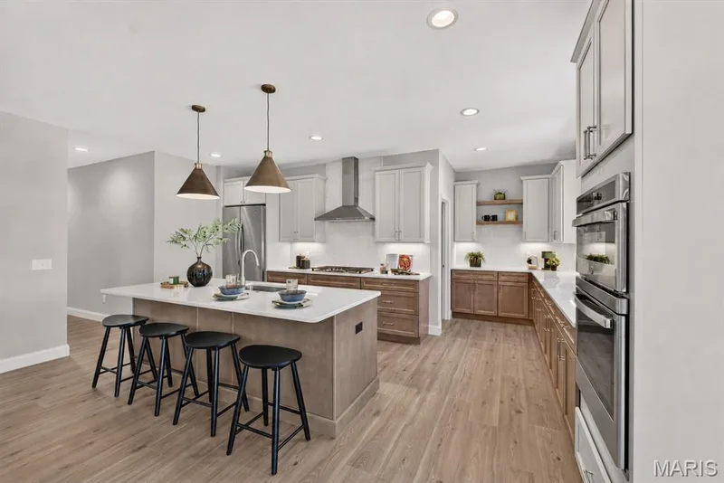 Open-concept kitchen featuring an oversized island with seating, pendant lighting, stainless steel appliances, dual-tone cabinetry,  wood-finish flooring, and walk-in pantry — 126 Royal Troon Drive, Dardenne Prairie, MO 63368