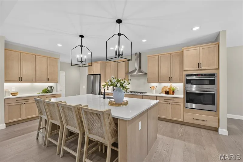 Spacious kitchen featuring an expansive central island with counter seating, light wood-finish cabinetry, stainless steel appliances, and two contemporary pendant lights — 128 Royal Troon Drive, Dardenne Prairie, MO 63368