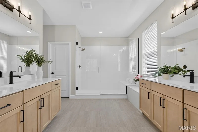 Dual wood-finish vanities with white countertops, matte black fixtures, walk-in shower with clear glass enclosure, and light-toned tile flooring — 128 Royal Troon Drive, Dardenne Prairie, MO 63368