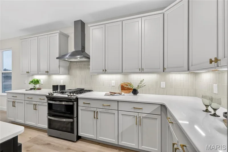 Modern kitchen featuring light gray cabinetry, a gas range with a stainless steel vent hood, white countertops, and a light-toned tile backsplash — 366 Strawberry Grove Drive, O'Fallon, MO 63376