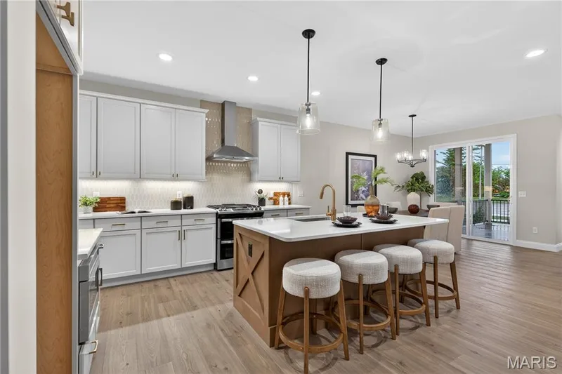 Open-concept kitchen featuring light wood-finish flooring, white cabinetry, a large kitchen island with seating, a stainless steel range hood, and a sliding glass door — 369 Strawberry Grove Drive, O'Fallon, MO 63376