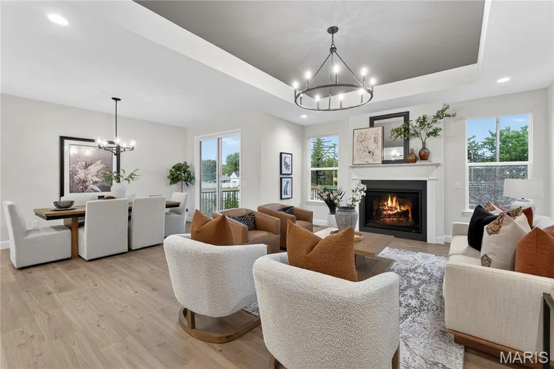 Open-concept living space featuring wood-finish flooring, a tray ceiling with a contemporary chandelier, a built-in fireplace with a mantel, and multiple windows providing natural light — 369 Strawberry Grove Drive, O'Fallon, MO 63376
