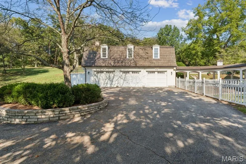Two-car garage with dormer windows, paved driveway, stone retaining wall, mature trees, and a white picket fence — 2333 Ossenfort Road, Wildwood, MO 63038