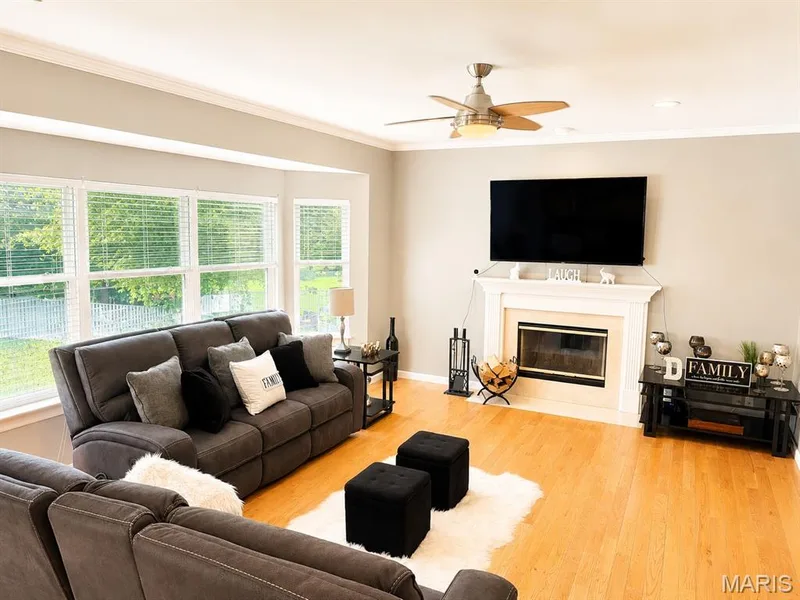 Bright living space featuring natural wood-finish flooring, a bay window overlooking exterior greenery, a built-in fireplace with a white mantel, recessed lighting, and a ceiling fan — 1126 Manor Cove Drive, St Charles, MO 63304