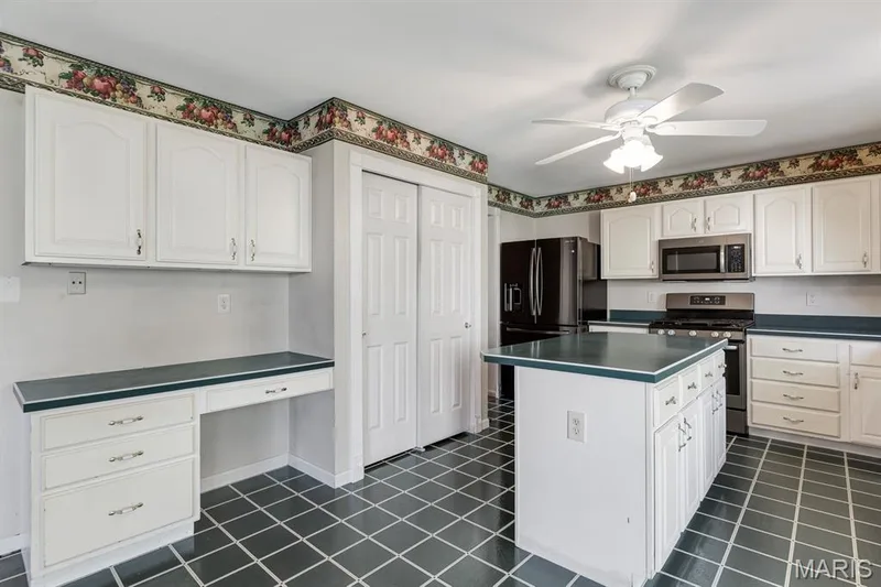 Spacious kitchen featuring an island, white cabinetry, dark countertops, stainless steel appliances, and dark square floor tiles — 1103 Panda Court, Wentzville, MO 63385