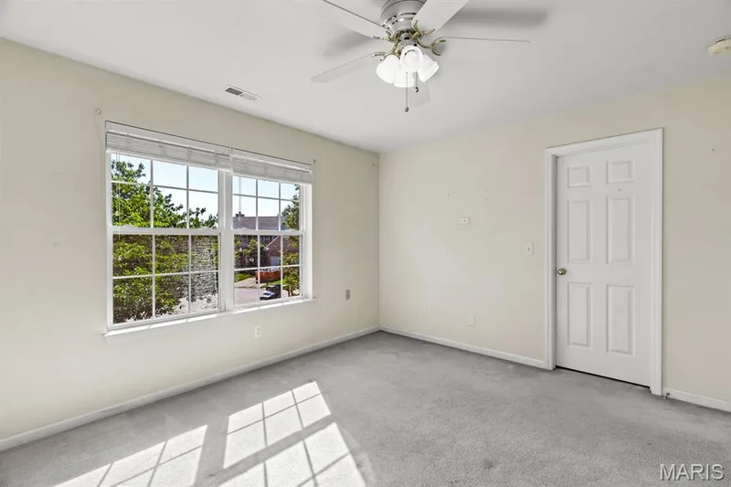 Bedroom featuring a large window with white grilles, light-colored walls, gray carpeting, a ceiling fan with integrated lighting, and a paneled door with a round knob — 1103 Panda Court, Wentzville, MO 63385