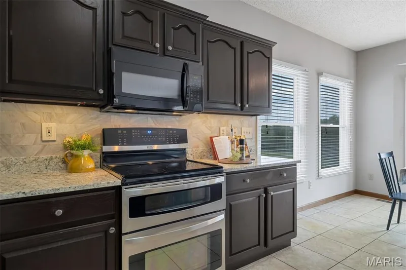 Kitchen featuring dark wood cabinetry with silver hardware, granite-style countertops, and a stone-look tile backsplash — 195 England Drive, O'Fallon, MO 63366