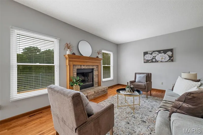 Living space featuring a brick fireplace with a wood mantel, light gray walls, wood-finish flooring, and white window blinds — 195 England Drive, O'Fallon, MO 63366