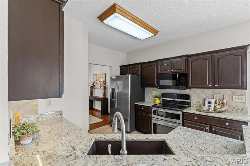Kitchen featuring dark wood cabinetry, light-toned granite countertops, stainless steel appliances, a built-in microwave, and a double basin sink with a gooseneck faucet — 195 England Drive, O'Fallon, MO 63366