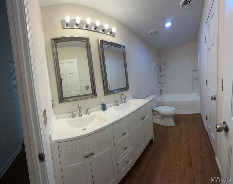 Dual vanity with integrated sinks, white cabinetry, wood-finish flooring, full bathtub with shower, and overhead lighting — 2505 Ambelwood Lane, O'Fallon, MO 63368