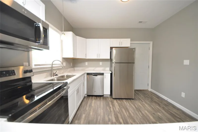 Kitchen featuring white shaker cabinetry, stainless steel appliances, light-toned countertops, wood-finish flooring, and a double basin sink — 502 S Linn Avenue, Wentzville, MO 63385