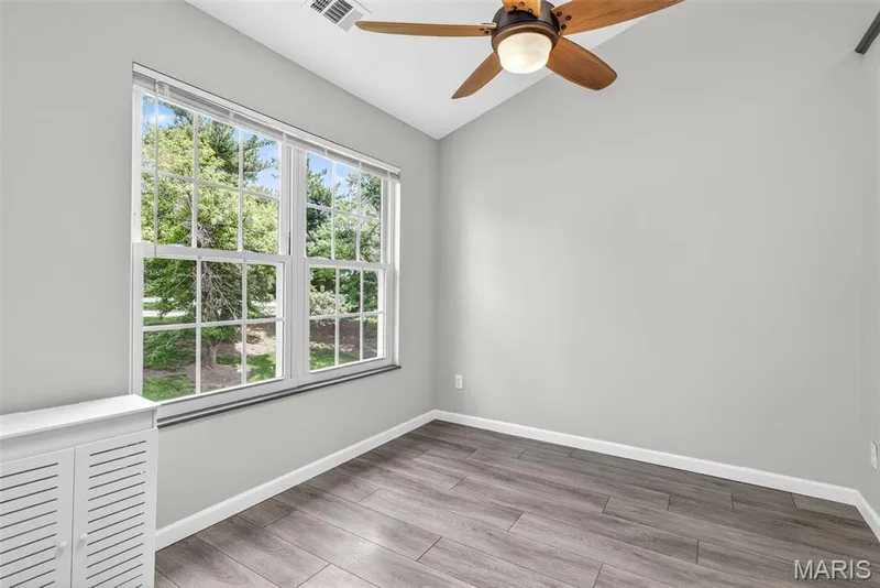 Bright room featuring a large double window, wood-finish flooring, a ceiling fan with integrated lighting, and neutral wall tones — 1034 Windstream Drive #14, St Peters, MO 63376