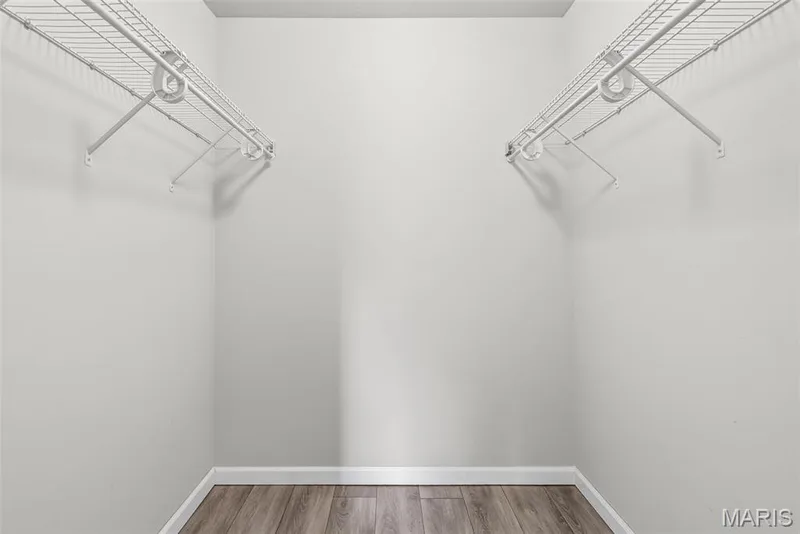 Walk-in closet featuring light gray walls, white baseboards, wood-finish flooring, and two white wire shelving units with hanging rods — 1034 Windstream Drive #14, St Peters, MO 63376