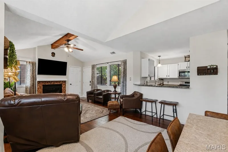 Living area featuring a vaulted ceiling with a wood beam, fireplace with mosaic tile surround, wood-finish flooring, and an open-concept kitchen with white cabinetry and a breakfast bar — 704 Hemingway Lane #E, Weldon Spring, MO 63304