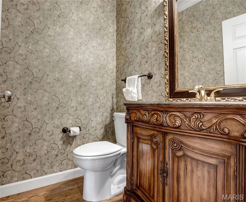 Main Floor ½ Bathroom. Decorative wallpaper, ornate wood-finish vanity with a stone countertop, framed mirror, and wood-finish flooring — 2154 White Lane Drive, Chesterfield, MO 63017