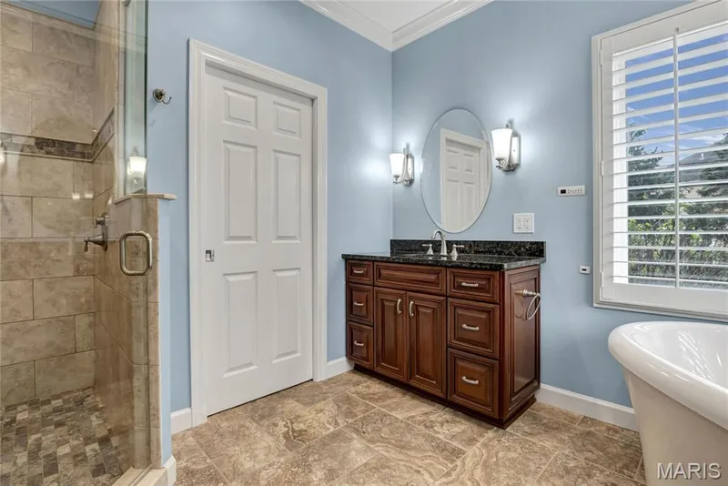 Tiled walk-in shower with glass enclosure, wood vanity with dark countertop, oval mirror, plantation shutters, and a freestanding bathtub — 2154 White Lane Drive, Chesterfield, MO 63017