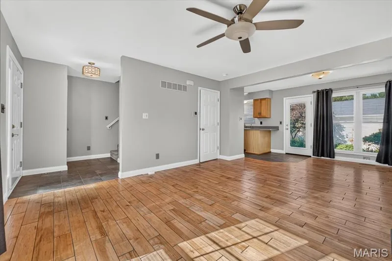 Open-concept living area featuring wood-finish flooring, a ceiling fan, and large windows with black curtains — 115 Carriage View Drive, Wildwood, MO 63040