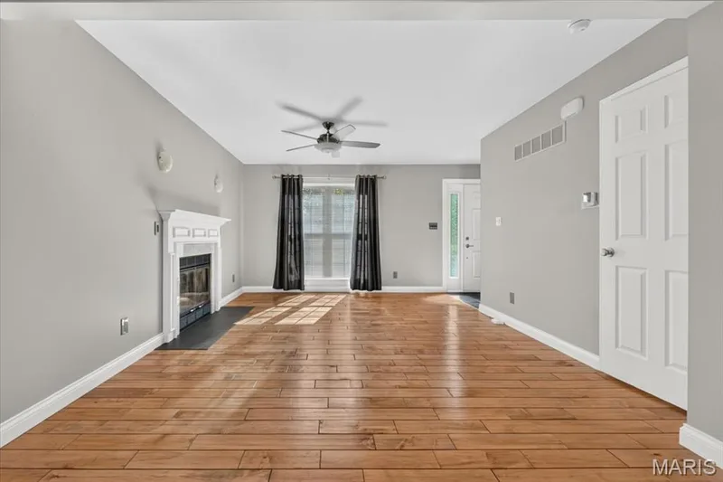 Spacious room featuring wood-finish flooring, a white mantel fireplace with a dark hearth, a ceiling fan, and large windows with dark drapery — 115 Carriage View Drive, Wildwood, MO 63040