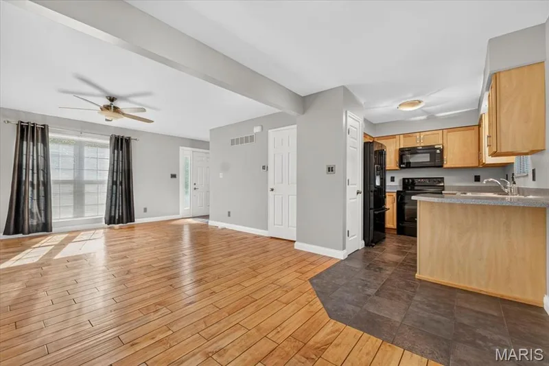 Open concept living area featuring wood-finish flooring, a ceiling fan, and a kitchen with light wood cabinetry, dark countertops, and black appliances — 115 Carriage View Drive, Wildwood, MO 63040