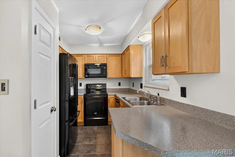 Kitchen featuring light wood cabinetry, black appliances, speckled countertops, and dark tile flooring — 115 Carriage View Drive, Wildwood, MO 63040