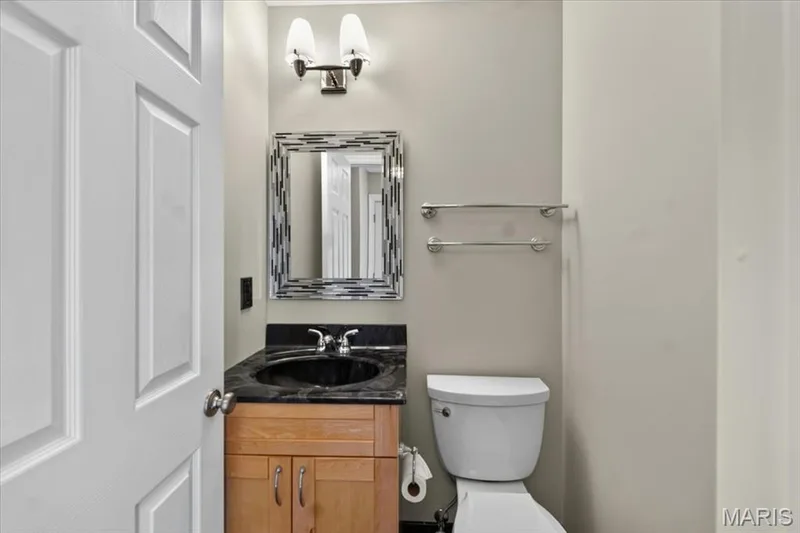 Bathroom featuring a wood-finish vanity with an integrated dark countertop and undermount sink, a toilet, a decorative framed mirror, and a wall-mounted light fixture — 115 Carriage View Drive, Wildwood, MO 63040