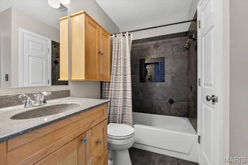 Bathroom featuring a granite-finish countertop with an integrated sink, natural wood cabinetry, and a shower-tub combination with dark-toned tile surround — 115 Carriage View Drive, Wildwood, MO 63040