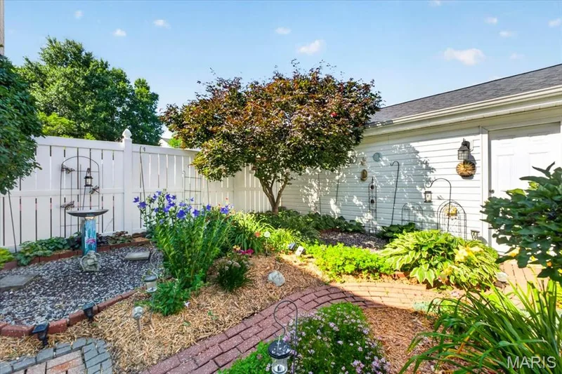 Landscaped garden area featuring mature tree, brick-edged planting beds, and white privacy fencing — 115 Carriage View Drive, Wildwood, MO 63040