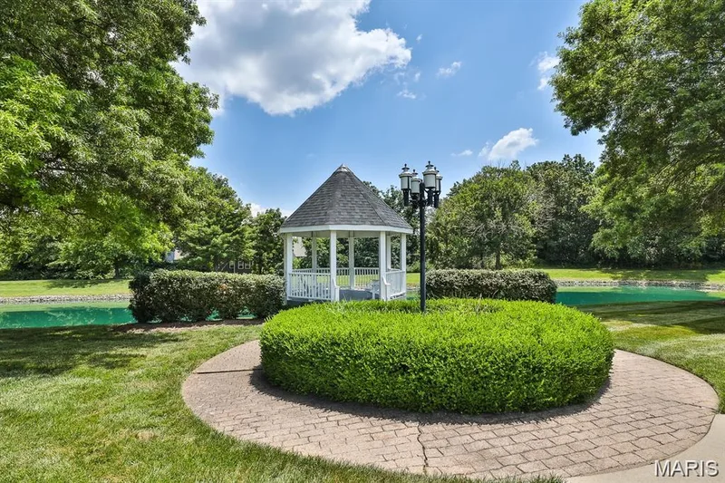 White gazebo with a dark shingle roof, surrounded by mature trees, lush green lawn, and a body of water — 115 Carriage View Drive, Wildwood, MO 63040