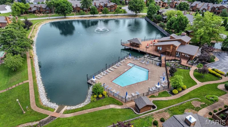 Community amenities featuring a central pond with a fountain, a large swimming pool, an expansive poolside deck, a multi-level clubhouse with exterior decking, and manicured green spaces with paved pathways — 309 Hemingway Lane, Weldon Spring, MO 63304