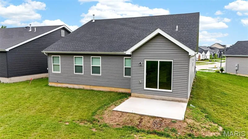 Rear exterior featuring gray horizontal siding, a dark shingle roof, three windows with white trim, and a sliding glass door opening to a concrete patio — 112 Creek Haven Court, Lake St. Louis, MO 63367