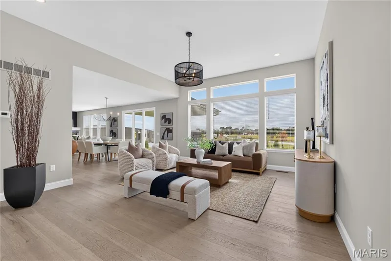 Expansive living area featuring light wood-finish flooring, a contemporary chandelier, and large windows — 26 Valor Way, O'Fallon, MO 63385