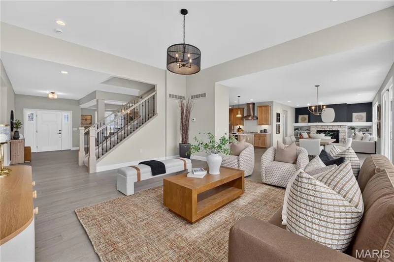 Spacious living area featuring wood-finish flooring, a contemporary chandelier, and a prominent staircase with wood railing — 26 Valor Way, O'Fallon, MO 63385