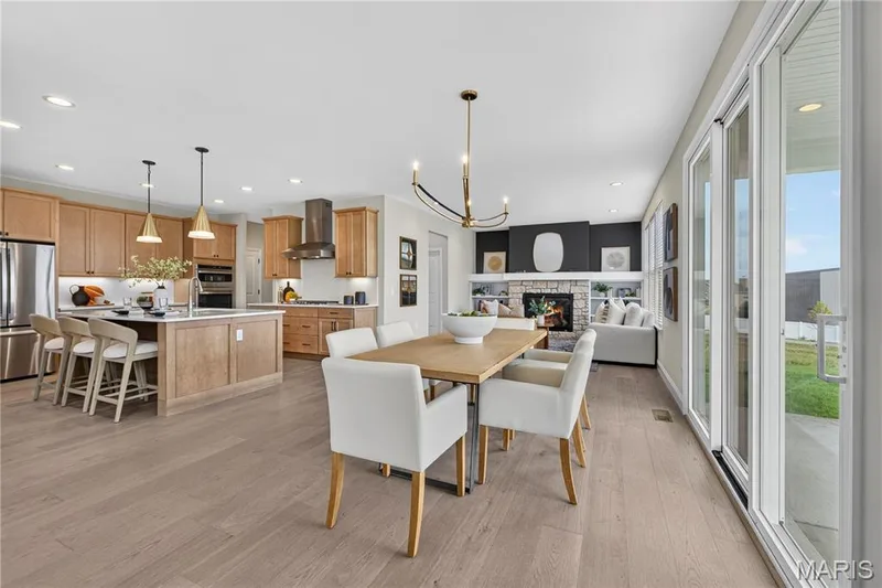 Open-concept living space featuring wood-finish flooring, a kitchen island with seating, a stone-surround fireplace, and large sliding glass doors — 26 Valor Way, O'Fallon, MO 63385