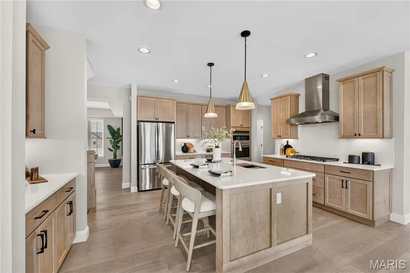 Spacious kitchen featuring an expansive center island, integrated stainless steel appliances, light wood-finish cabinetry, recessed lighting, and wood-finish flooring — 26 Valor Way, O'Fallon, MO 63385