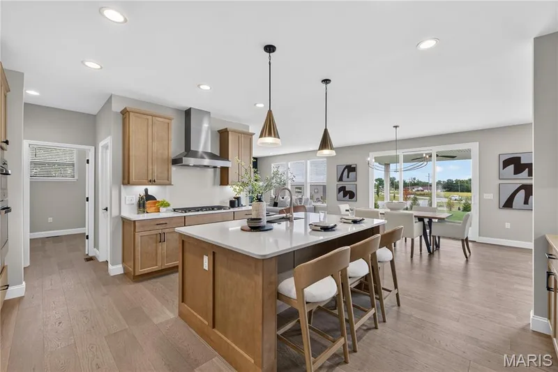 Recessed lighting illuminates the kitchen, featuring light wood-finish cabinetry, a large kitchen island with a white countertop, and wood-finish flooring throughout — 26 Valor Way, O'Fallon, MO 63385
