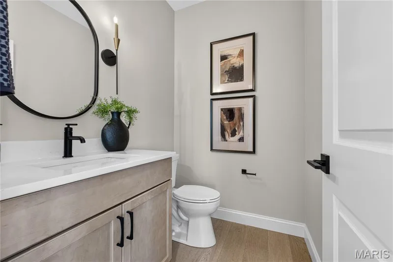 Main floor powder bathroom featuring light wood-finish flooring, a contemporary vanity with a white countertop, and a black-framed oval mirror — 26 Valor Way, O'Fallon, MO 63385