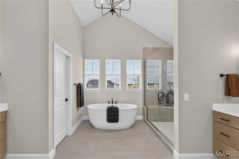 Vaulted ceiling with an open chandelier, freestanding soaking tub, tiled walk-in shower with a glass enclosure, and two wood-finish vanities with white countertops — 26 Valor Way, O'Fallon, MO 63385