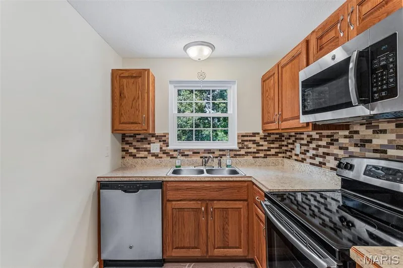 Kitchen featuring wood-finish cabinetry, stainless steel appliances, a mosaic tile backsplash, and a double basin sink — 1511 Hampton Hall Drive #10, Chesterfield, MO 63017
